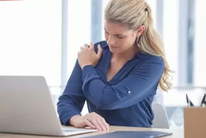 Businesswoman sitting at a desk with a laptop, holding her shoulder in pain, illustrating symptoms of shoulder pain caused by tendonitis or bursitis.
