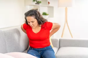 Image of a woman in a red shirt sitting on a couch, holding her lower back in pain, illustrating discomfort from lower back pain at home.